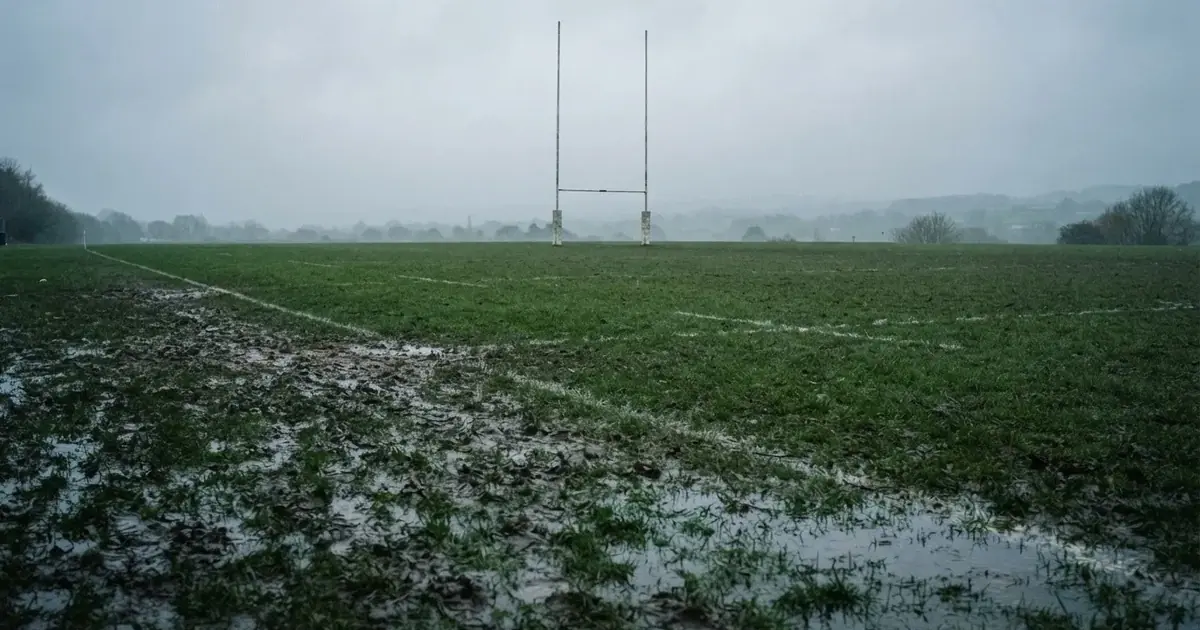 Boisko do rugby ze słupami bramki w oddali pod pochmurnym niebem, mokra trawa i ślady butów.